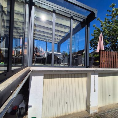 Glasveranda mit Holzdeck und Sonnenschirm, strahlender blauer Himmel im Hintergrund.