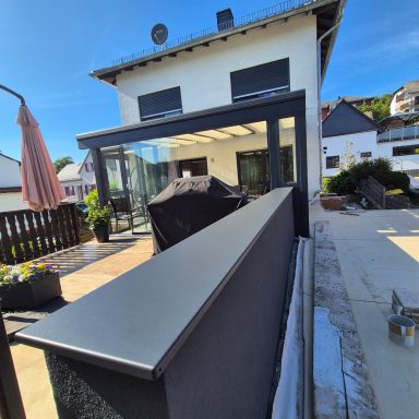 Moderne Terrassenansicht mit einem Grill und überdachtem Sitzbereich hinter einem Haus.